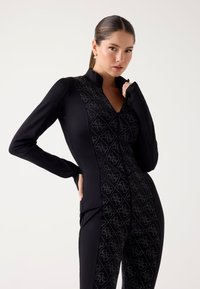 Czarny kombinezon z długim rękawem z zamkiem błyskawicznym z przodu, teksturowaną wstawką w diamentowy wzór oraz dopasowanym krojem. Wykonany z elastycznego materiału.