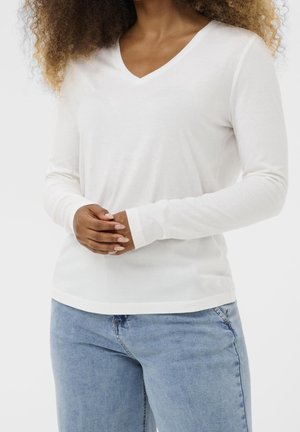 Femme portant un t-shirt blanc à manches longues avec col en V et un jean bleu clair, debout avec les mains doucement jointes devant elle.