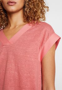 Růžový top s výstřihem do V a krátkými rukávy s ohrnutím, vyrobený z lehkého, texturovaného materiálu. Výstřih a okraje rukávů mají mírně tmavší růžový akcent.