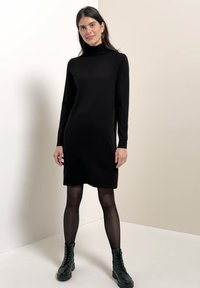 Frau trägt ein schwarzes, langärmliges Rollkragenkleid, schwarze transparente Strumpfhosen und schwarze geschnürte Knöchelstiefel und steht in einem neutralen Umfeld.