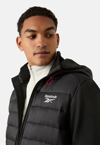 Czarna kurtka puchowa z kapturem, z pikowaną fakturą, srebrnym logo Reebok oraz czerwonymi akcentami. Nosi się ją na białej bluzie na zamek.