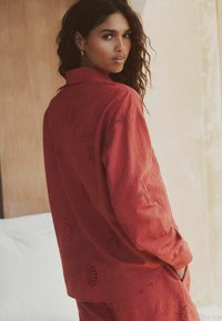 Punane linane särk lõigatud detailidega; pikad varrukad, nööbitavad mansetid ja kaarduvhem. Kandmiseks koos sobivate pükstega, pehme tekstuur, vabaaja stiil.