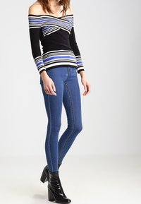 Frau trägt einen gestreiften Pullover mit schulterfreiem Schnitt in Schwarz, Weiß und Blau, enge blaue Jeans und schwarze Lackleder-Stiefeletten.