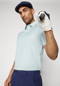 Light blue polo shirt with subtle texture, paired with navy pants. The subject holds a golf club with a white glove on one hand.