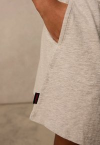 Ljust grå plagg med en texturerad yta, utrustad med en stor sidoficka och en svart etikett med "Superdry" i röd färg.