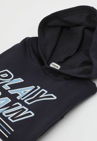 Granatowy sweatshirt z kapturem z kieszenią kangurką i puszystym wnętrzem. Z przodu znajduje się wyraźny napis "PLAY" w niebieskim i białym kolorze, z poziomymi liniami poniżej.