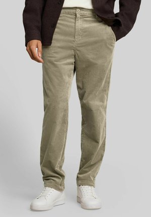 Mann trägt hellbraune Cordhosen, weiße Sneakers und eine dunkle Jacke mit einer Hand in der Tasche vor grauem Hintergrund.