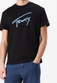 Černé bavlněné tričko s krátkými rukávy, na kterém je modré stylizované logo "Tommy Jeans" přes hruď. Nosí se s modrými džíny.
