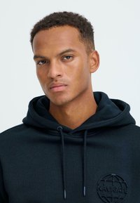 Felpa nera con cappuccio, con una patch logo testurizzata, cordini regolabili e un tessuto morbido e liscio. Vista ravvicinata della parte superiore del corpo.