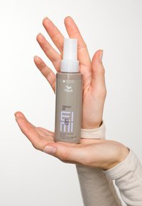 Grå plastflaska med en vit sprejmunstycke, märkt "Perfect Me" av Wella Professionals. Hållen i två händer med ljus hy.
