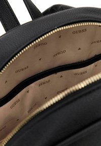 Sac à dos noir avec un extérieur texturé, une fermeture éclair dorée et un intérieur beige. L'intérieur présente un motif logo "GUESS" et une poche zippée.