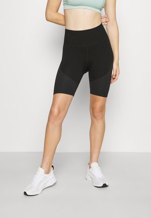 Kobieta ubrana w czarne, wysokie spodenki rowerowe, jasnografitowy top sportowy i białe sportowe buty, stojąca na jasnym, jednolitym tle.