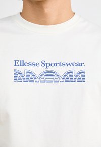 Balta medvilnė marškinėliai su mėlynų grafinių atspaudų „Ellesse Sportswear“, su arkos raštais ir horizontaliomis juostelėmis po jais.