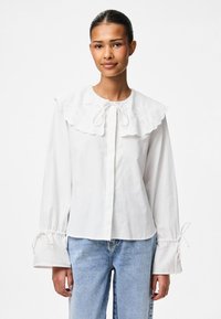 Weiße Bluse mit großem besticktem Kragen, Bindegürtel am Ausschnitt und elastischen Ärmelbündchen mit Kordelzug. Kombiniert mit hellblauen Jeans.
