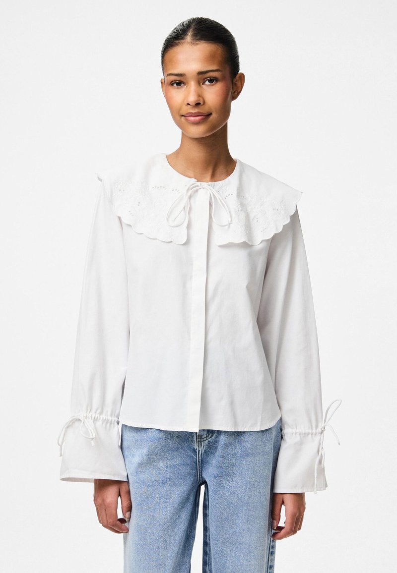 Weiße Bluse mit großem besticktem Kragen, Bindegürtel am Ausschnitt und elastischen Ärmelbündchen mit Kordelzug. Kombiniert mit hellblauen Jeans.