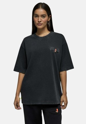 Femme aux longs cheveux foncés portant un t-shirt ample gris foncé et un pantalon noir avec de petits détails orange et beige sur les poches.