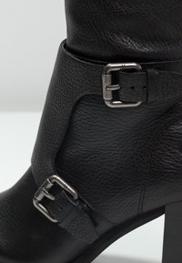 Musta nahkainen nilkkasaapas, jossa on teksturoitu pinta. Varustettu kahdella metallibucklilla ja jämäkällä korolla lisävakauden vuoksi.