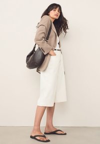Lys beige blazer, hvide shorts med vide ben, sort bælte og flip-flops. Model poserer med en sort læder skuldertaske mod en neutral baggrund.