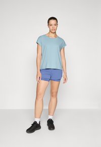 Camisa de manga corta azul claro combinada con pantalones cortos morados. El modelo lleva zapatillas negras y calcetines blancos, de pie contra un fondo neutro.