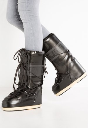 Personne portant des bottes d'hiver brillantes de style Moon Boot, noires, avec des semelles épaisses et des lacets, associées à un jean skinny gris clair sur fond blanc.