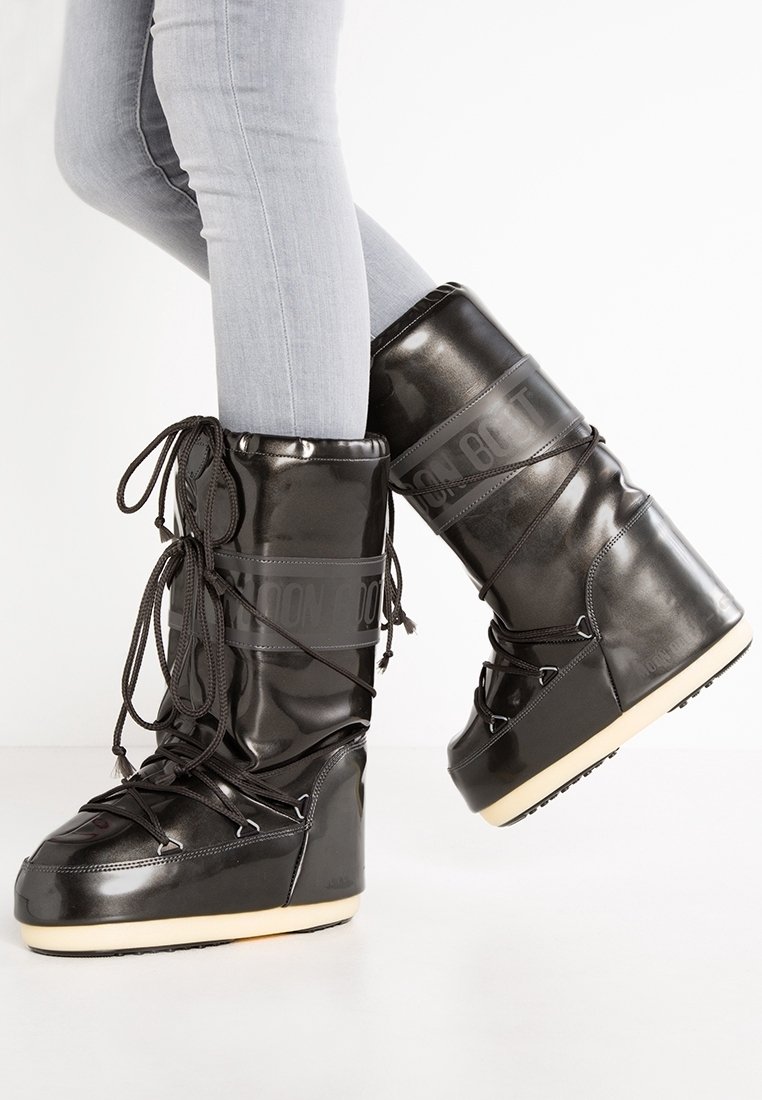 Personne portant des bottes d'hiver brillantes de style Moon Boot, noires, avec des semelles épaisses et des lacets, associées à un jean skinny gris clair sur fond blanc.