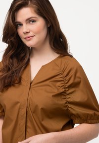 Brun bluse med pufærmer og V-hals, med samlet detaljering i siden og en glat, let stoftekstur.