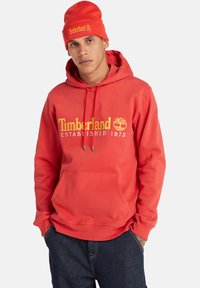 Hombre con sudadera roja de Timberland y gorro a juego, con las manos en los bolsillos, frente a un fondo claro y sencillo.