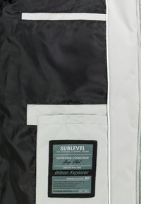 Schwarz-weißes Outerwear-Jackeninnere mit einem großen Etikett mit dem Markennamen "SUBLEVEL", Taschendetails und Nähakzenten.