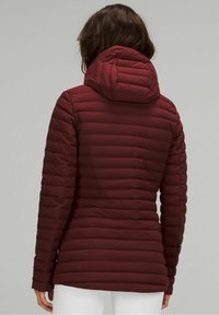 Kobieta w dopasowanej, bordowej pikowanej kurtce z kapturem, pokazana od tyłu, w białych spodniach na jednolitym tle.