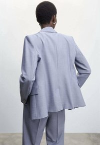 Blazer grigio chiaro su misura con una vestibilità rilassata, spalle strutturate, schiena allungata e tasche laterali. Tessuto liscio, dettagli minimi.