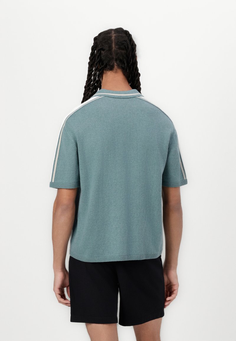 Polo à manches courtes, col, de couleur bleu canard avec une finition texturée et des rayures blanches sur les côtés. Associé à un short noir.