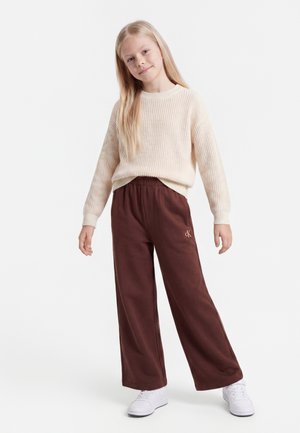 Jeune fille aux longs cheveux blonds portant un pull crème, un pantalon large marron et des baskets blanches, debout devant un fond blanc.
