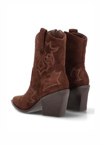 Botas vaqueras de ante marrón con tacón apilado, con patrones de costura decorativos en marrón más claro en los lados.