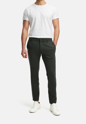 Homme portant un pantalon noir ajusté, un t-shirt blanc à col rond et des baskets blanches, debout avec une main dans la poche.