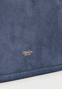 Μπλε σουέντ υλικό με χρυσό λογότυπο "Coach New York" ανάγλυφο πάνω από περαστή άκρη σε ανοιχτό φόντο.