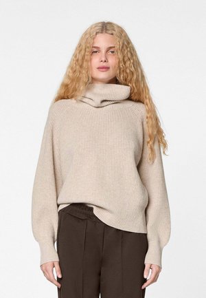 Frau mit langen lockigen blonden Haaren, die einen beige gerippten Rollkragenpullover und dunkelbraune Hosen trägt, steht vor einem schlichten weißen Hintergrund.