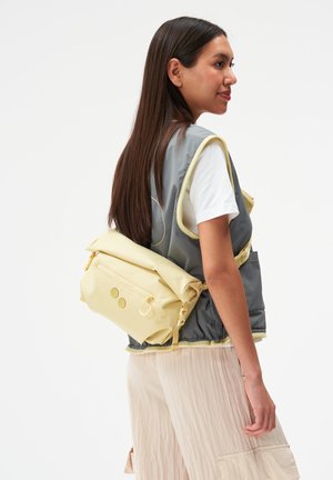 Femme aux longs cheveux bruns portant un gilet gris, une chemise blanche et un pantalon beige, portant un sac banane jaune clair.