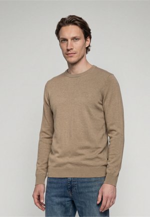 Homme aux cheveux courts et bruns portant un pull beige à manches longues et un jean bleu, debout devant un fond uni gris clair.