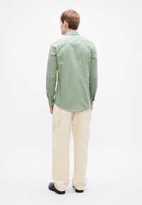 Homem de costas, vestindo uma camisa de manga longa verde-claro, calças largas creme com bolsos e sapatos pretos, em fundo branco.