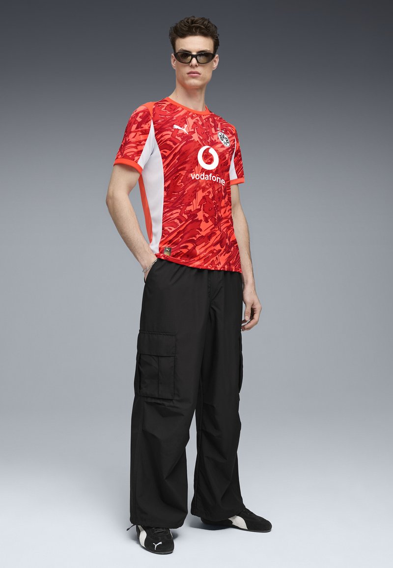 Rød mønstret trøje med hvide detaljer, der har Puma-logo og Vodafone-branding. Kombineret med sorte cargo-bukser og sorte sko.