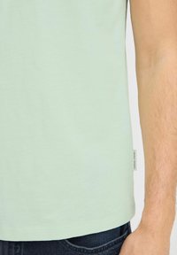 Svijetlo zeleni T-shirt s glatkom teksturom, s lagano zakrivljenim rubom i malom oznakom brenda sa strane. Nositi s tamnoplavim trapericama.