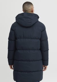 Polstret mørk marineblå parka med hette, med en glatt tekstur og horisontalt quiltet design. Den går til midt på låret, har lange ermer og skjulte lommer.