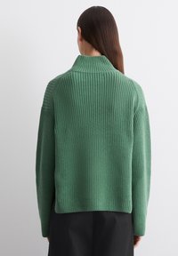 Grüner Strickpullover mit hohem Kragen, geripptem Muster und überschnittenen Schultern. Der Rücken ist leicht oversized und fällt unter die Taille.
