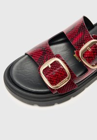 Rød sandal med skjellmønster i reptilskinn med to brede stropper og gullfargede spenner, og med en teksturert svart såle for grep og komfort.