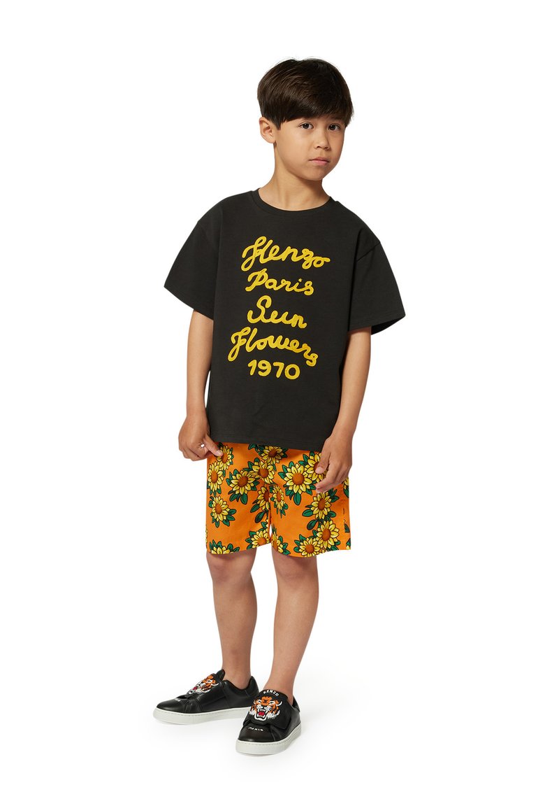 Czarna, oversize koszulka z żółtym napisem, pomarańczowe szorty w zielony wzór słoneczników oraz czarne trampki z wzorem w tigra.