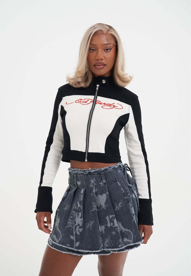 Ung kvinde med blond hår iført en sort og hvid cropped jakke med "Ed Hardy" tekst og en plisseret denimskørt med falmede grafiske tryk.