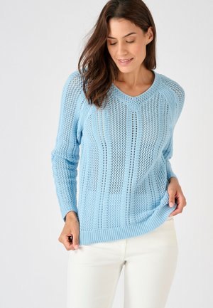 Donna con maglione a maglia azzurro chiaro a motivi verticali, abbinato a pantaloni bianchi, davanti a uno sfondo chiaro e uniforme.