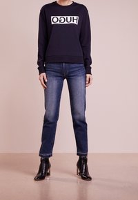 Granatowa bluza z kapturem z białym logo "HUGO" na przodzie, w połączeniu z niebieskimi dżinsami i czarnymi botkami na słupku.