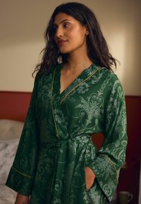 Robe verde con motivo floreale e maniche ampie, dotata di una vita con cintura e passamaneria gialla lungo il colletto e i polsini. Tessuto dalla superficie liscia.