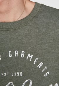 Sudadera verde oliva con un acabado texturizado. Presenta un cuello redondo y un estampado frontal con texto blanco que incluye "GARMENTS" y "EST. 1390."